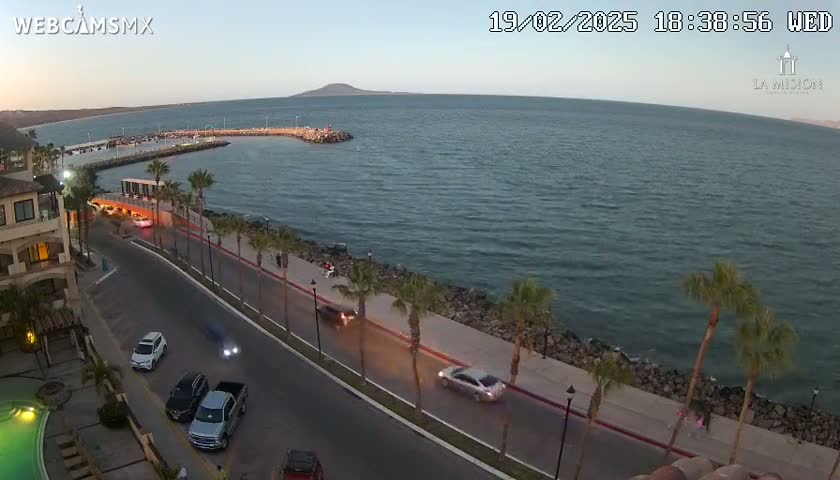 An oceanfront road with palm trees and cars driving along a paved walkway at dusk, overlooking a calm bay and distant mountains under a clear sky.