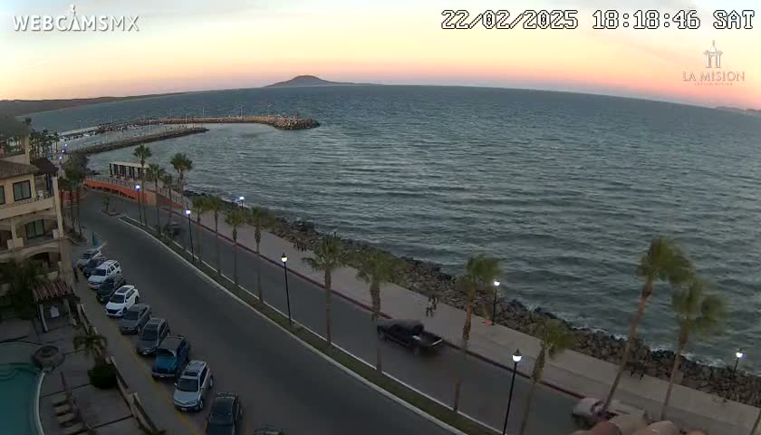 An aerial view at dusk shows a calm bay with a paved waterfront road lined with palm trees and cars parked along the side, next to a building.