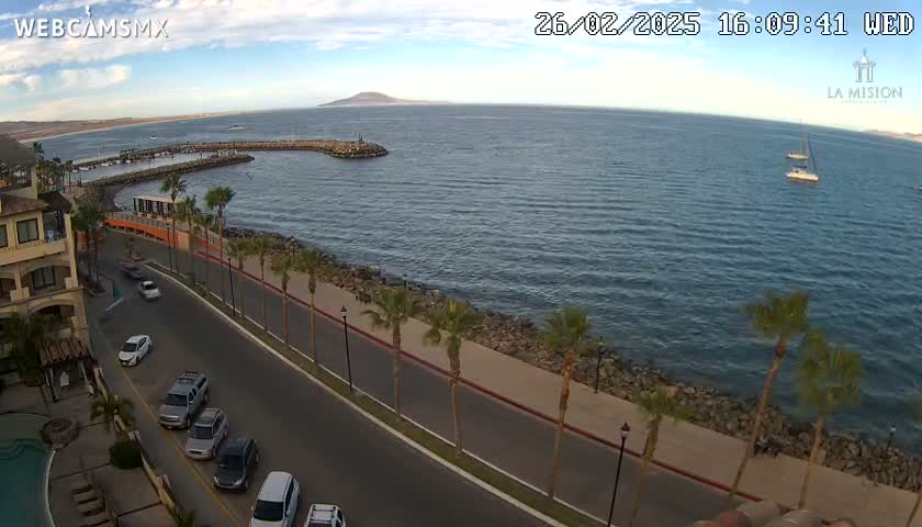 An oceanside road with palm trees and parked cars lines a calm bay under a partly cloudy sky, with several sailboats visible in the distance and a distant island.