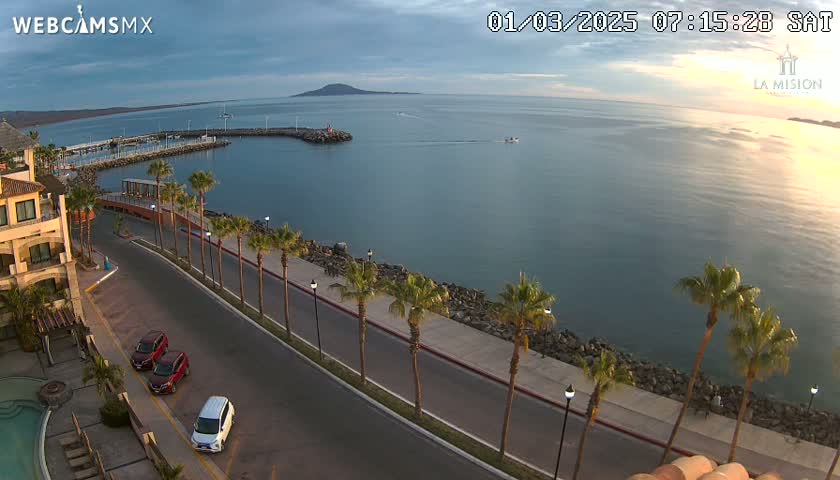 A coastal road lined with palm trees curves alongside a calm bay, under a partly cloudy sky, with a few vehicles parked along the roadside.
