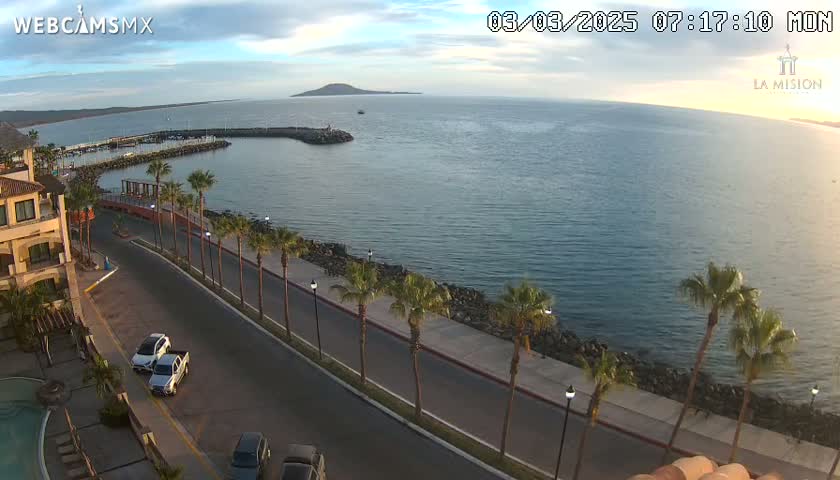 A partly cloudy morning scene shows a calm ocean, a marina, a palm tree-lined road next to a building, and several parked cars.