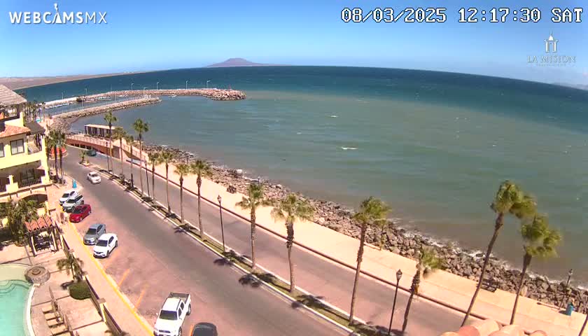 A sunny day overlooks a coastal road lined with palm trees, leading to a calm ocean with a small harbor, and a distant mountain visible on the horizon.