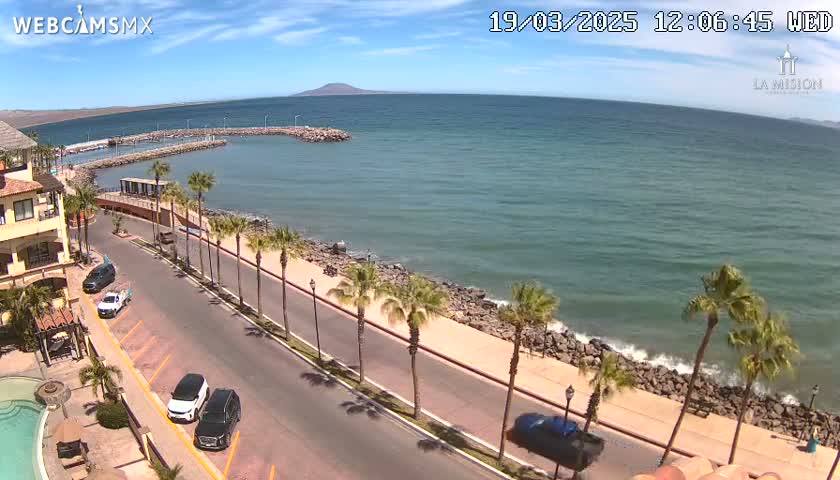 A seaside road lined with palm trees curves along a rocky coastline with calm, blue-green water under a mostly sunny sky.