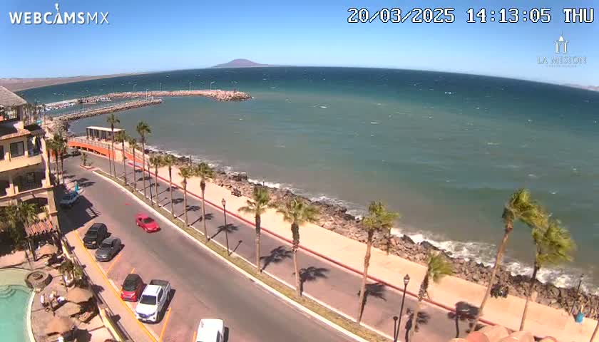 A sunny, windy day at a coastal town shows a road lined with palm trees alongside a calm ocean with a small harbor and distant mountains.