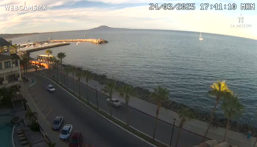 An aerial view shows a calm ocean with a small boat and a distant island at sunset, overlooking a seaside road with palm trees and parked cars.