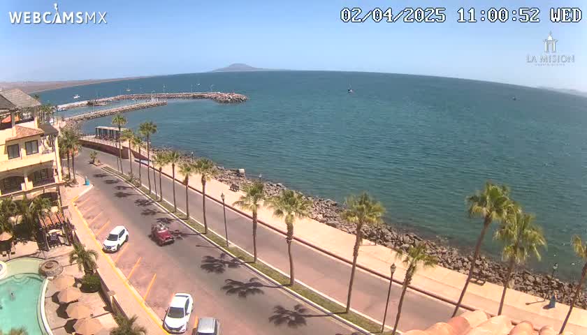 A sunny day overlooking a coastal road lined with palm trees, leading to a calm ocean with a small island in the distance.