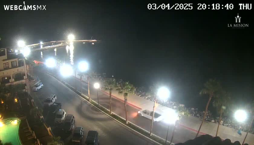 A nighttime, overhead view shows a coastal road with parked cars and a few moving vehicles near a body of water under bright artificial lights.