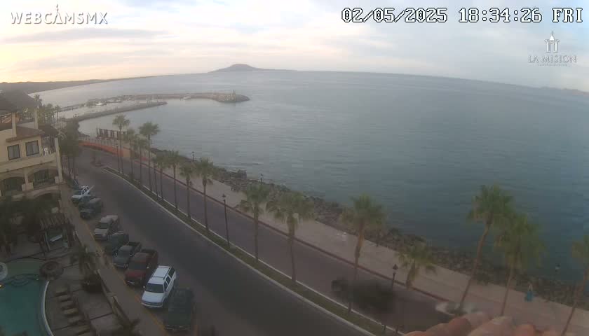 An aerial view of a coastal town at dusk shows a calm ocean, a palm-tree lined road, and parked cars near a building with a swimming pool.