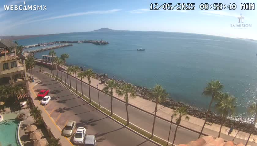 A coastal road lined with palm trees overlooks a calm, blue ocean with a small boat and a distant island under a mostly sunny sky.