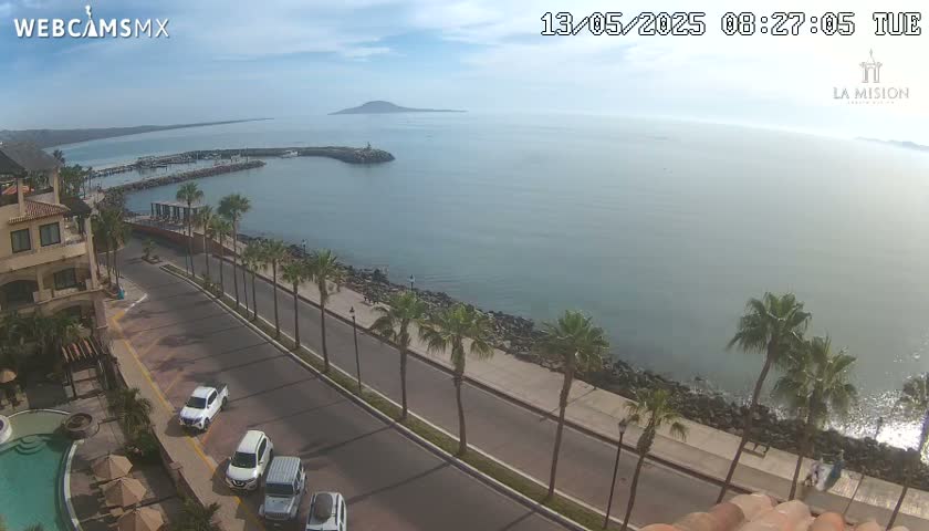 A partly sunny seaside road with palm trees lines a paved walkway next to a calm ocean with a small island in the distance.