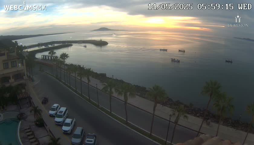A sunrise over calm ocean water, viewed from an elevated position, shows a road, palm trees, parked cars, and a few boats in the distance.
