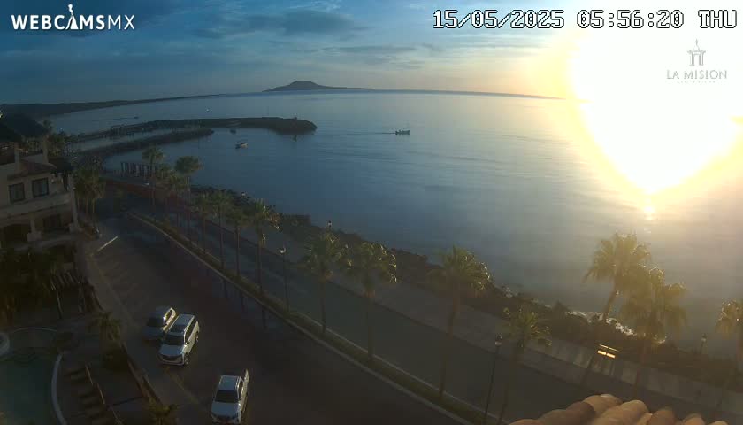 A sunrise over a calm ocean, viewed from an elevated position overlooking a coastal road lined with palm trees and a few parked cars.