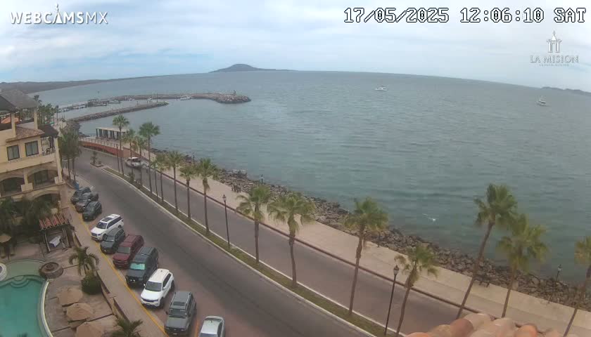 An elevated view shows a calm ocean harbor with a road lined with palm trees leading to a building next to a swimming pool under a partly cloudy sky.