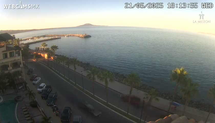 An elevated view of a calm ocean harbor at sunset, with a coastal road lined with palm trees and parked cars in front of a building.