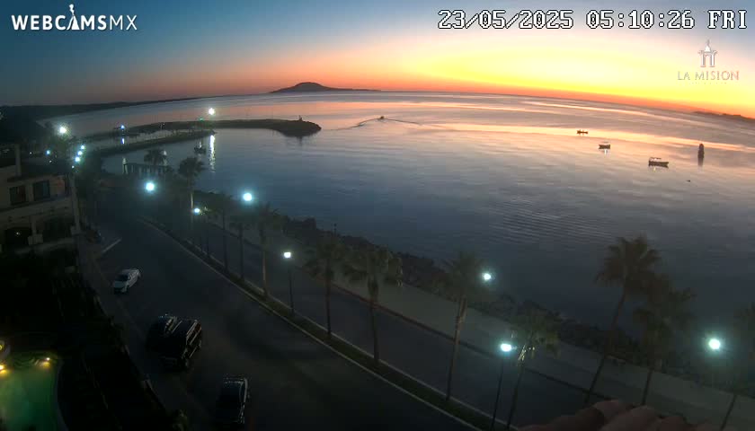 An elevated view shows a calm body of water at sunrise, with a coastal road lined with palm trees and streetlights, and several boats visible in the bay.