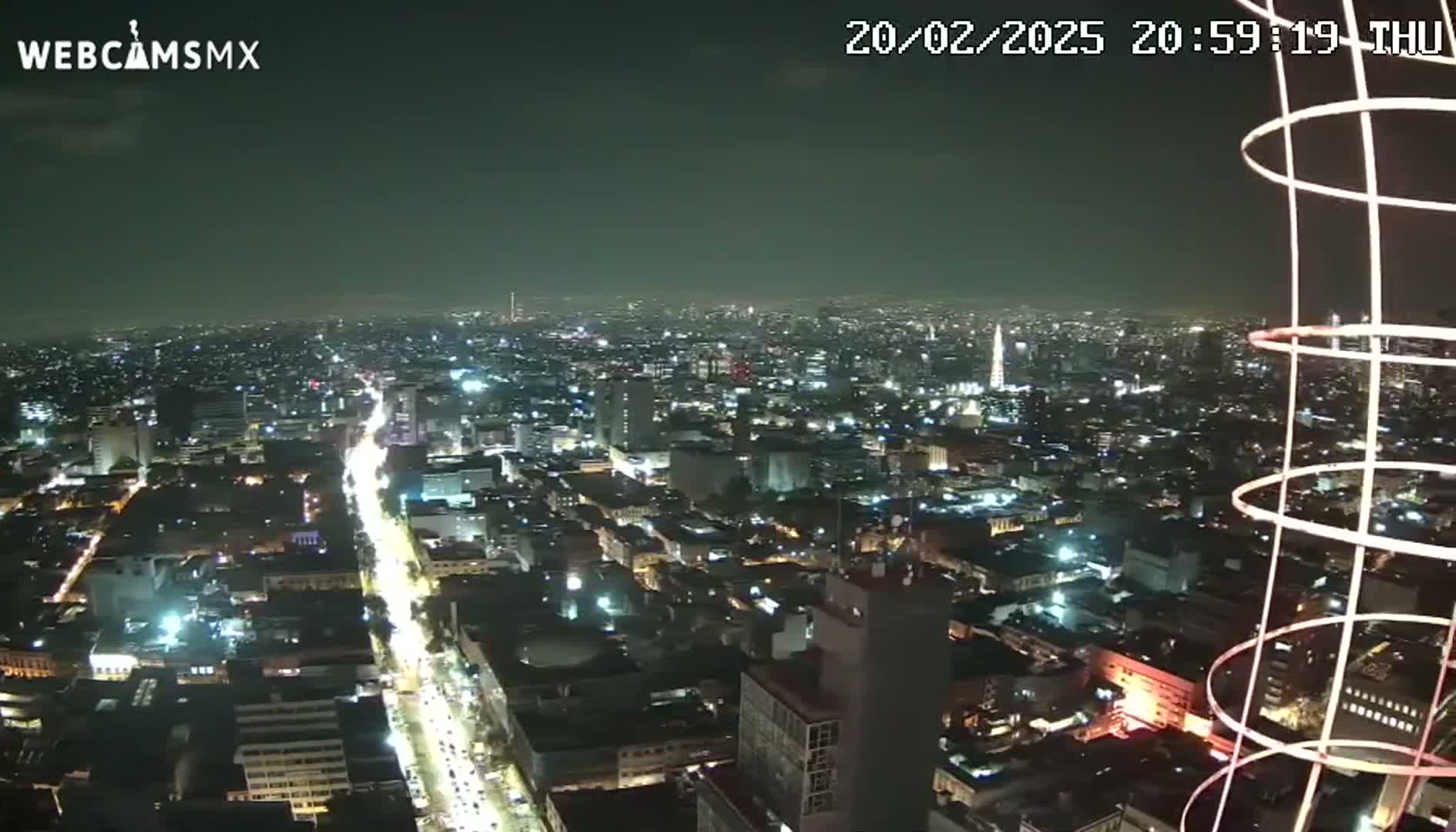 A nighttime, high-angle view of a sprawling city illuminated by streetlights and building lights, partially obscured by a tall, circular structure in the foreground.