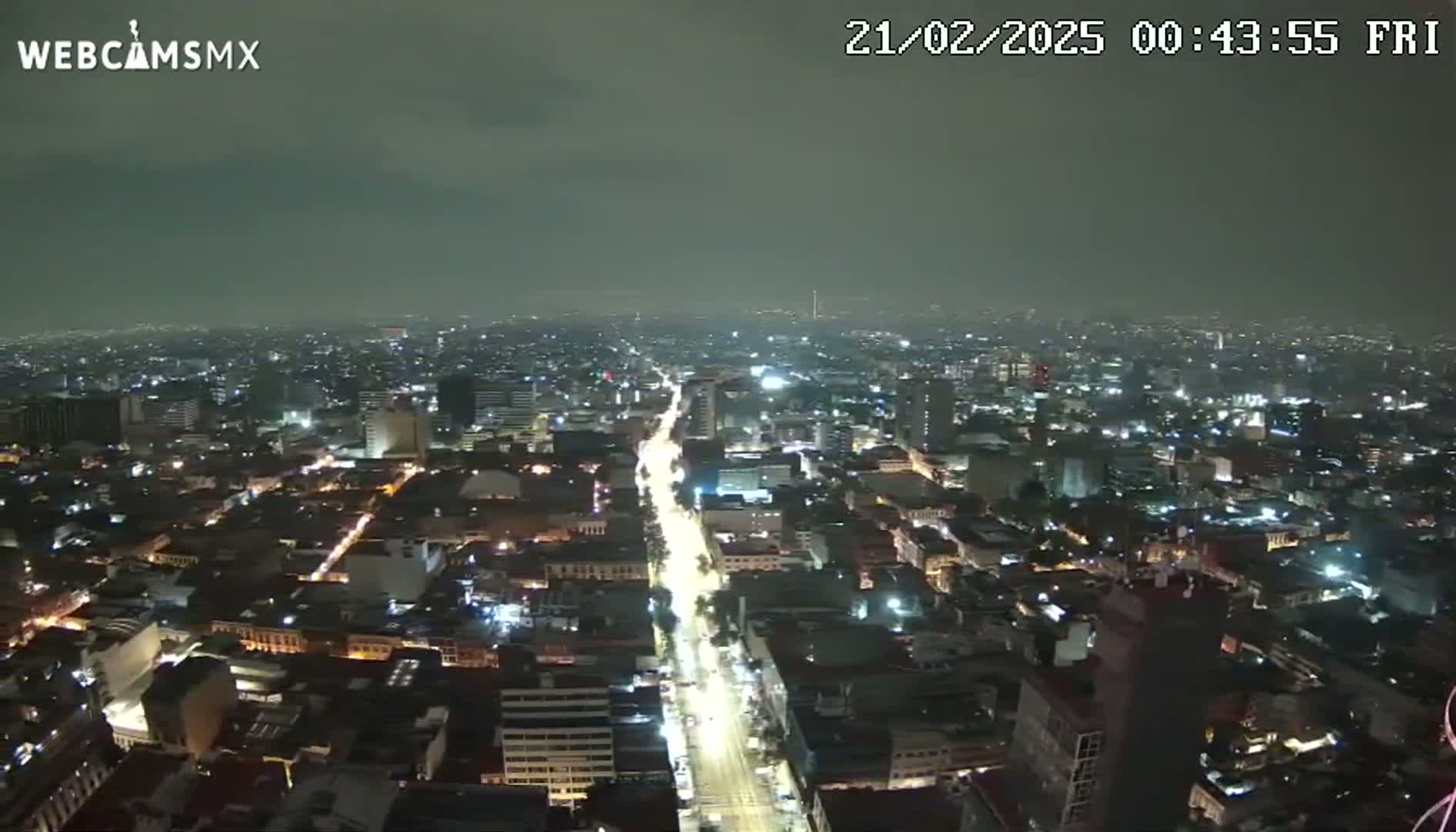 A nighttime, high-angle view of a sprawling city with overcast skies shows a brightly lit main street cutting through the densely packed buildings.