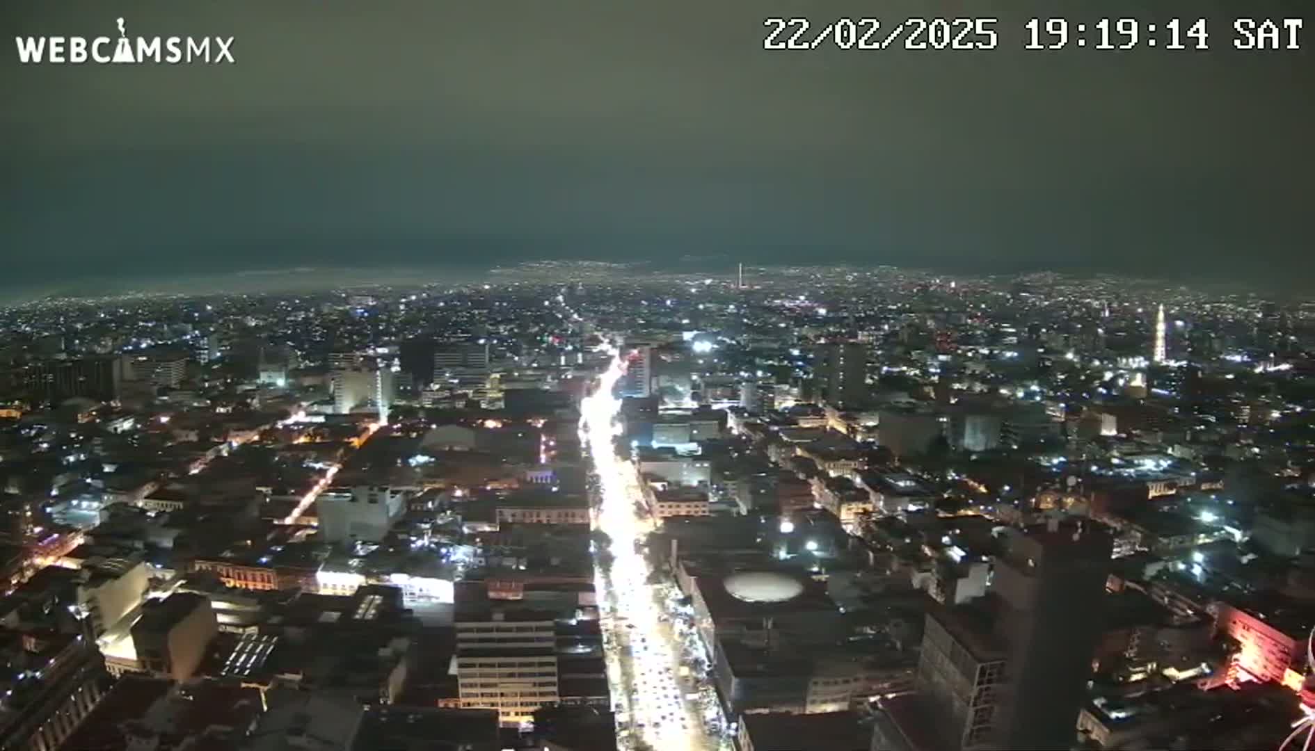 A nighttime aerial view of a city with a hazy sky shows a long, brightly lit street cutting through the densely packed buildings.
