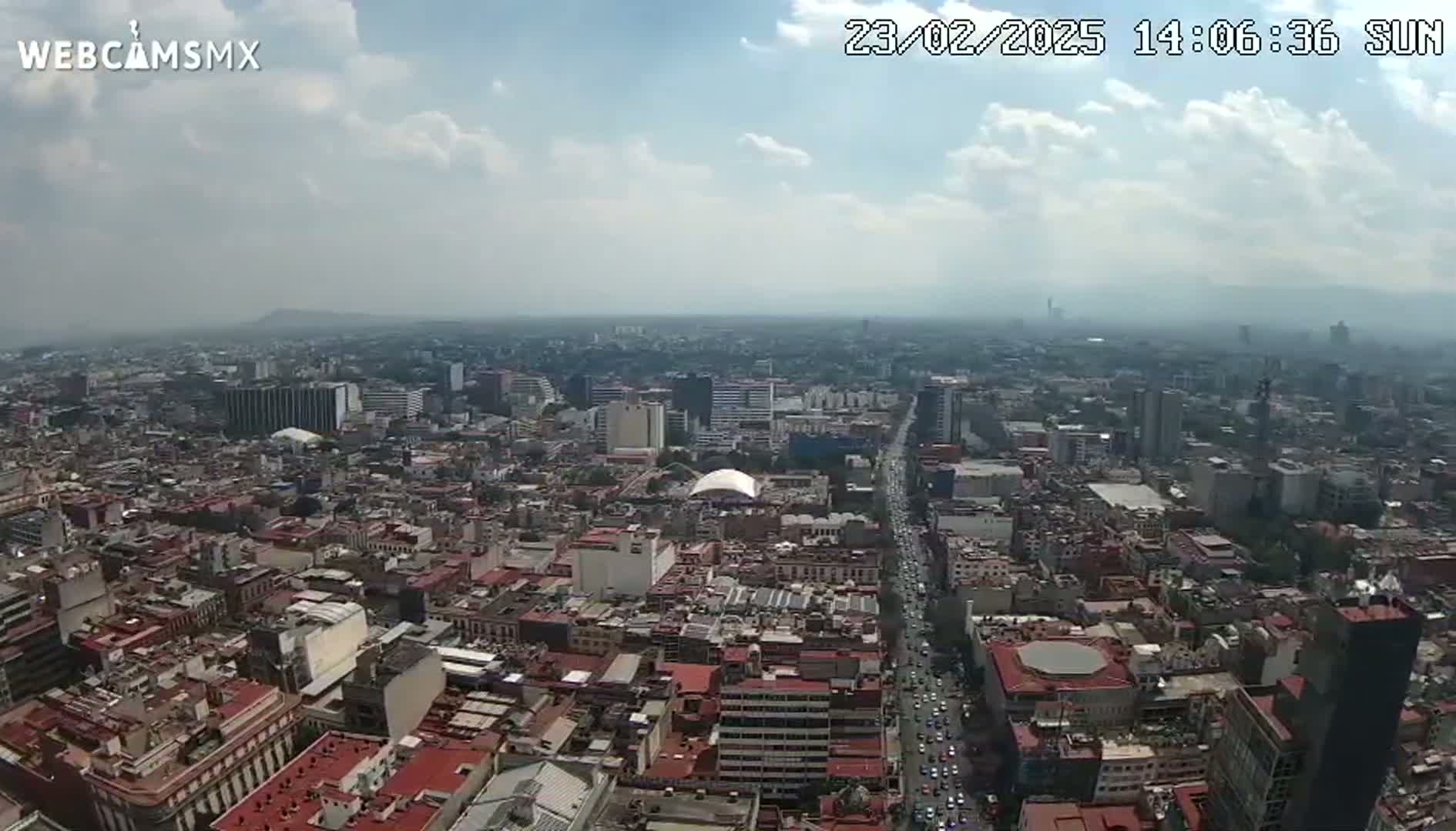 A high-angle, long shot of a sprawling city on a partly cloudy day shows numerous buildings and streets extending to the horizon.