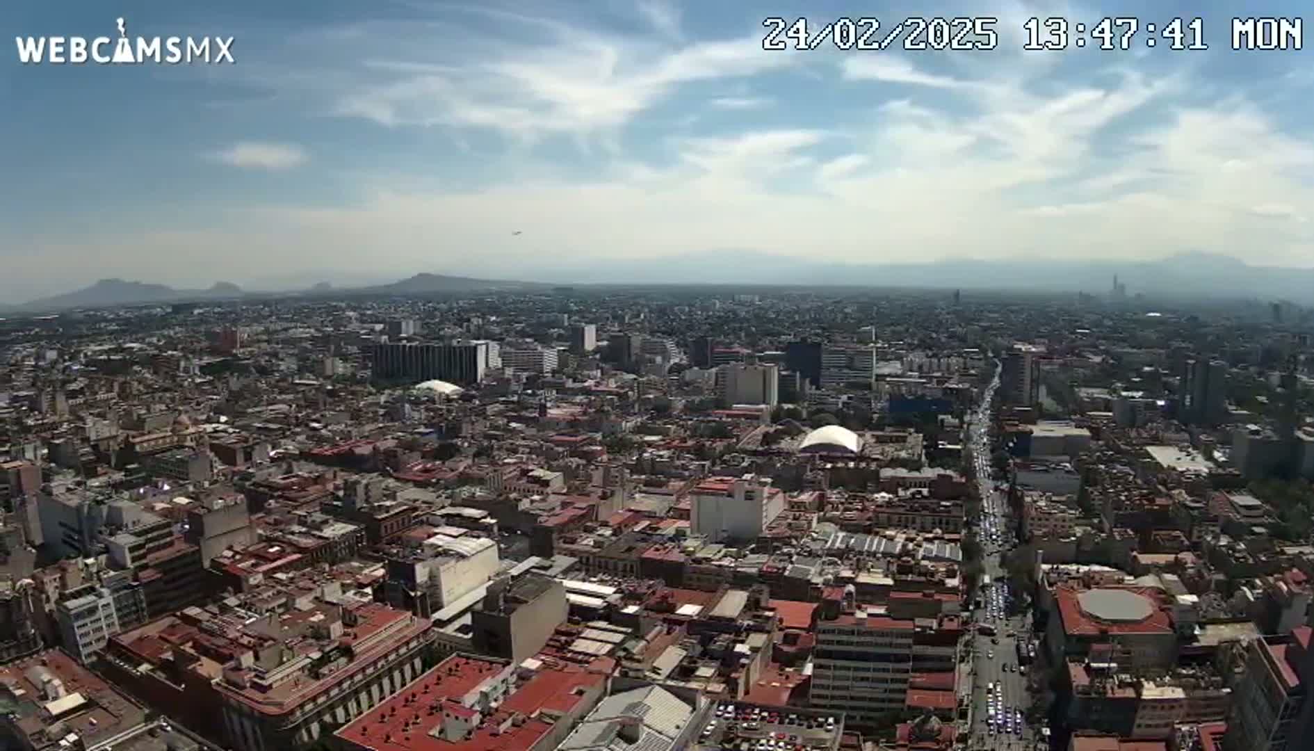 A high-angle, long shot of a sprawling city under a partly cloudy sky shows distant mountains on the horizon.