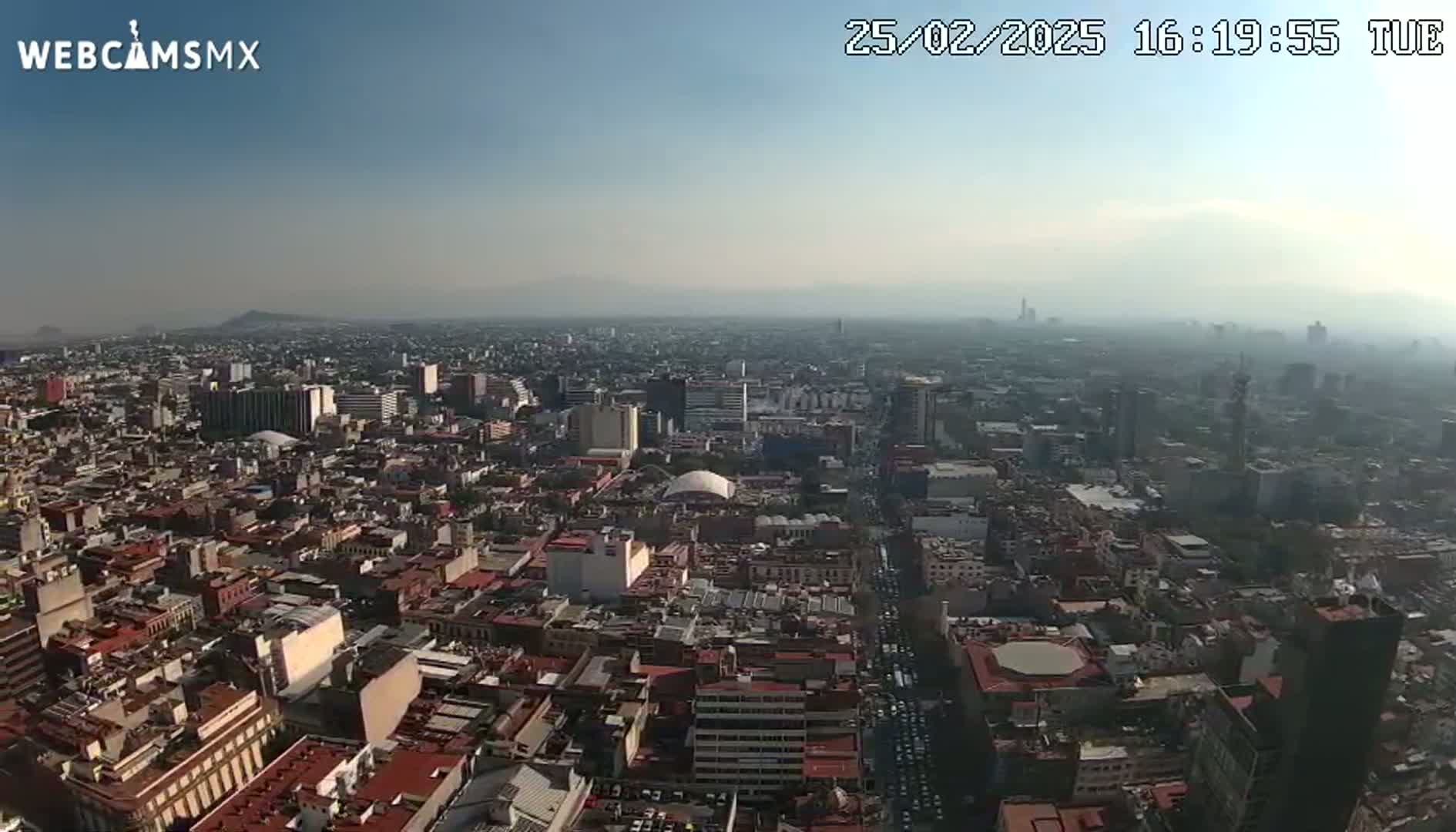 A hazy, sunny day reveals a sprawling cityscape, viewed from a high vantage point, showing densely packed buildings extending to a distant mountain range.