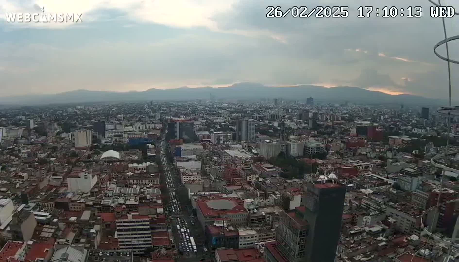 A high-angle, long shot of a sprawling city under a partly cloudy, dusky sky shows distant mountains on the horizon.