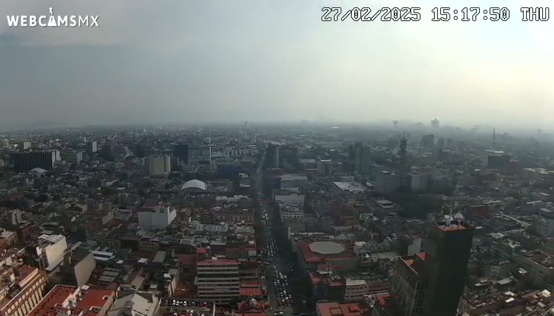 A hazy, overcast day reveals a sprawling cityscape viewed from a high vantage point, showing numerous buildings and streets extending to the horizon.