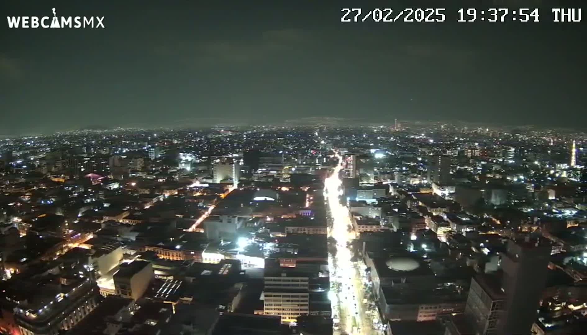 A nighttime, high-angle view shows a sprawling city illuminated by countless lights, with a major thoroughfare brightly lit as it cuts through the urban landscape.
