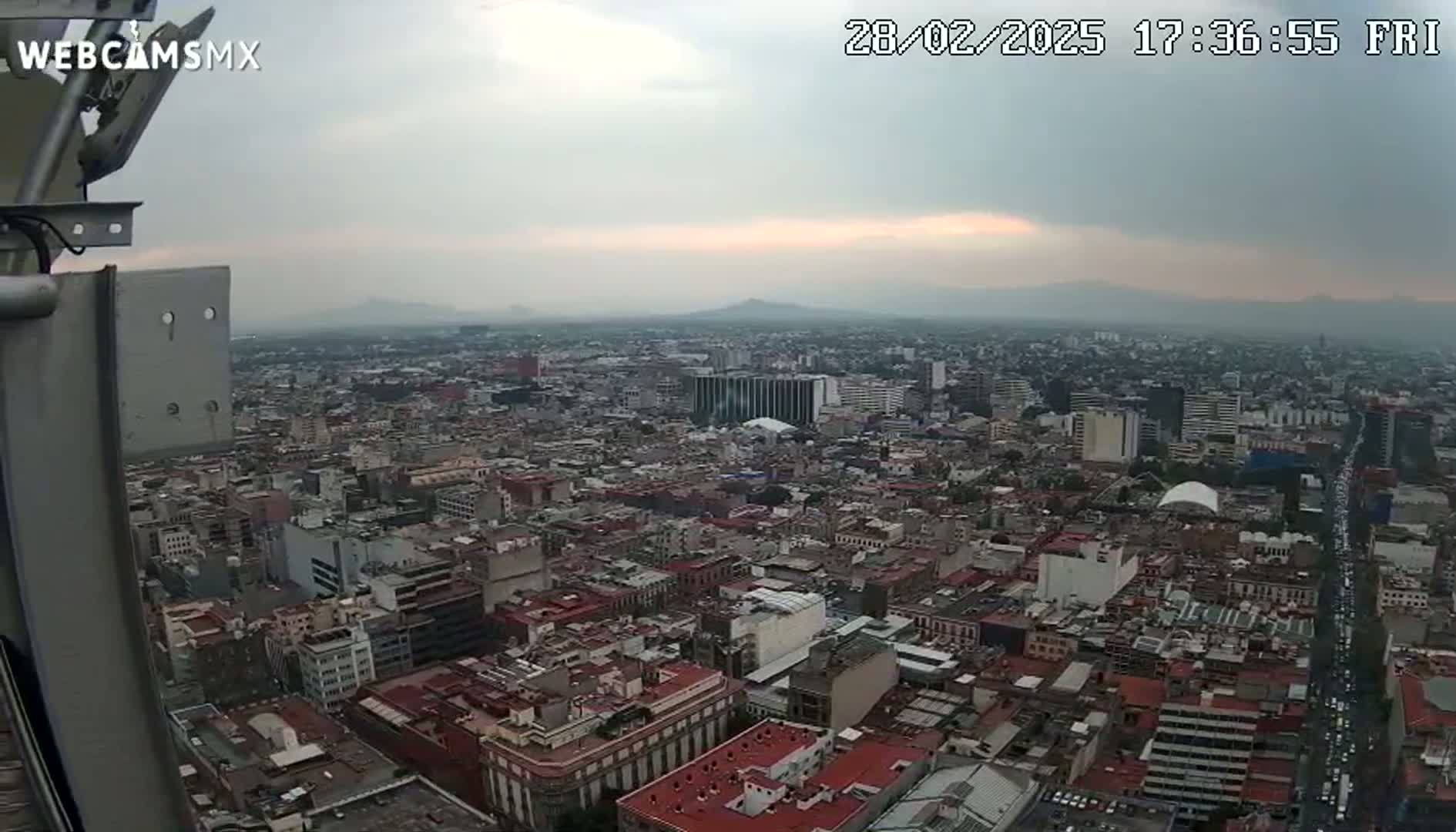 A high-angle, long shot of a sprawling city under a cloudy, overcast sky shows a vast expanse of buildings and streets stretching to distant mountains on the horizon.