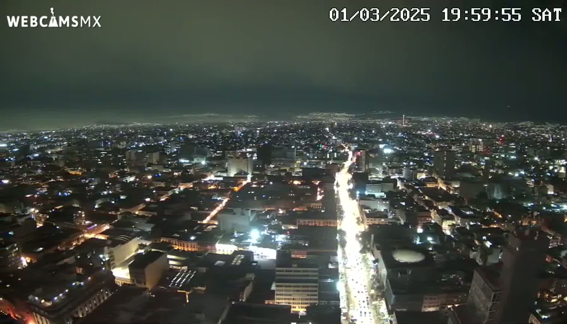 A nighttime aerial view of a city with a main street brightly lit, extending from the foreground into the distance under a mostly clear sky.