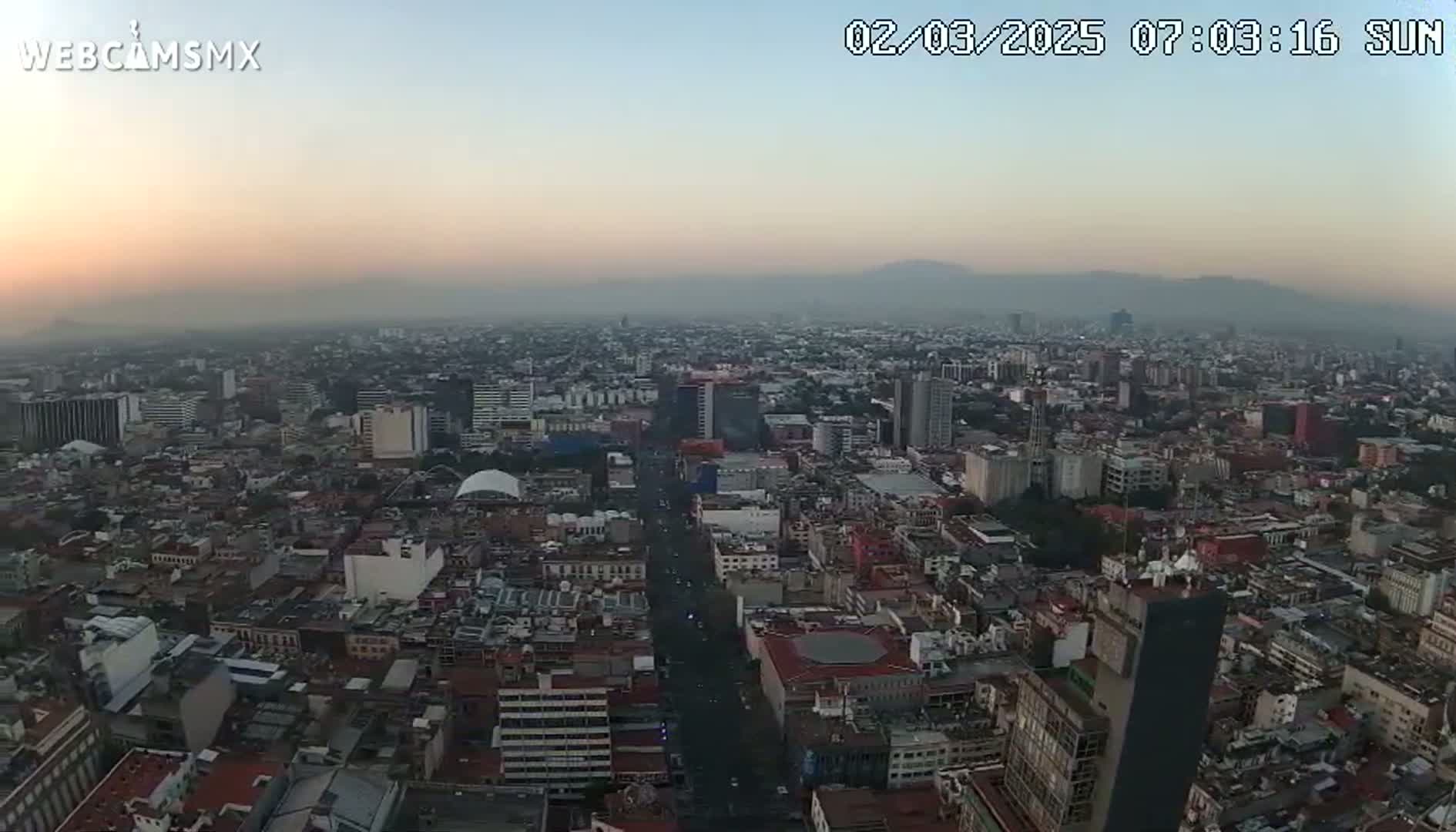 An aerial view of a hazy city at sunrise, showing a vast expanse of buildings and streets extending to distant mountains.