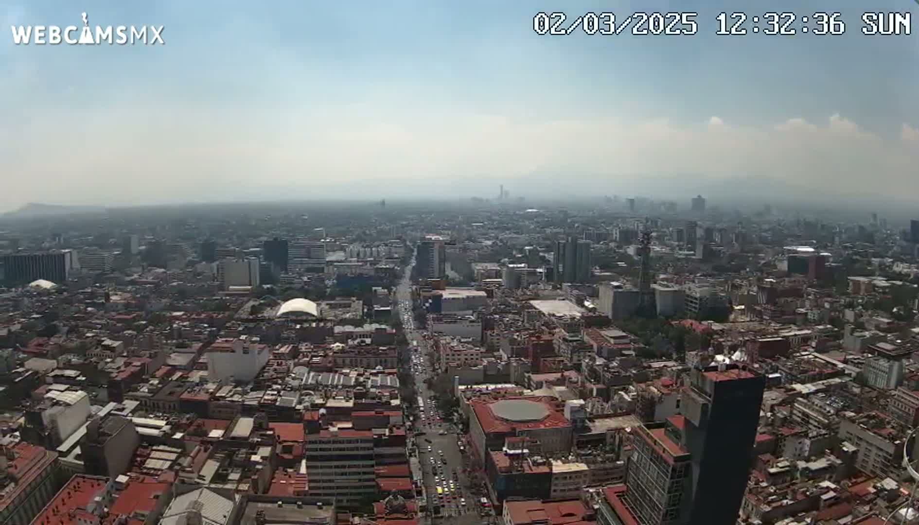 A hazy, sunny day reveals a sprawling cityscape viewed from a high vantage point, showing dense urban development extending to the horizon.