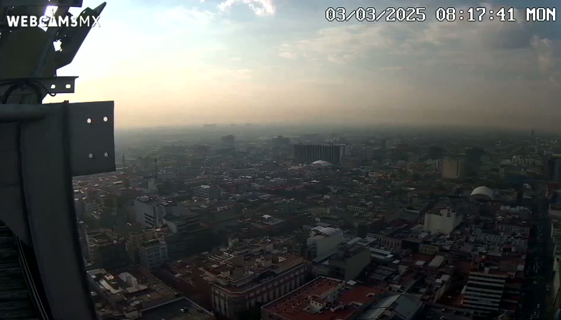 A hazy, partly cloudy morning view from a tall structure shows a sprawling city extending to the horizon.