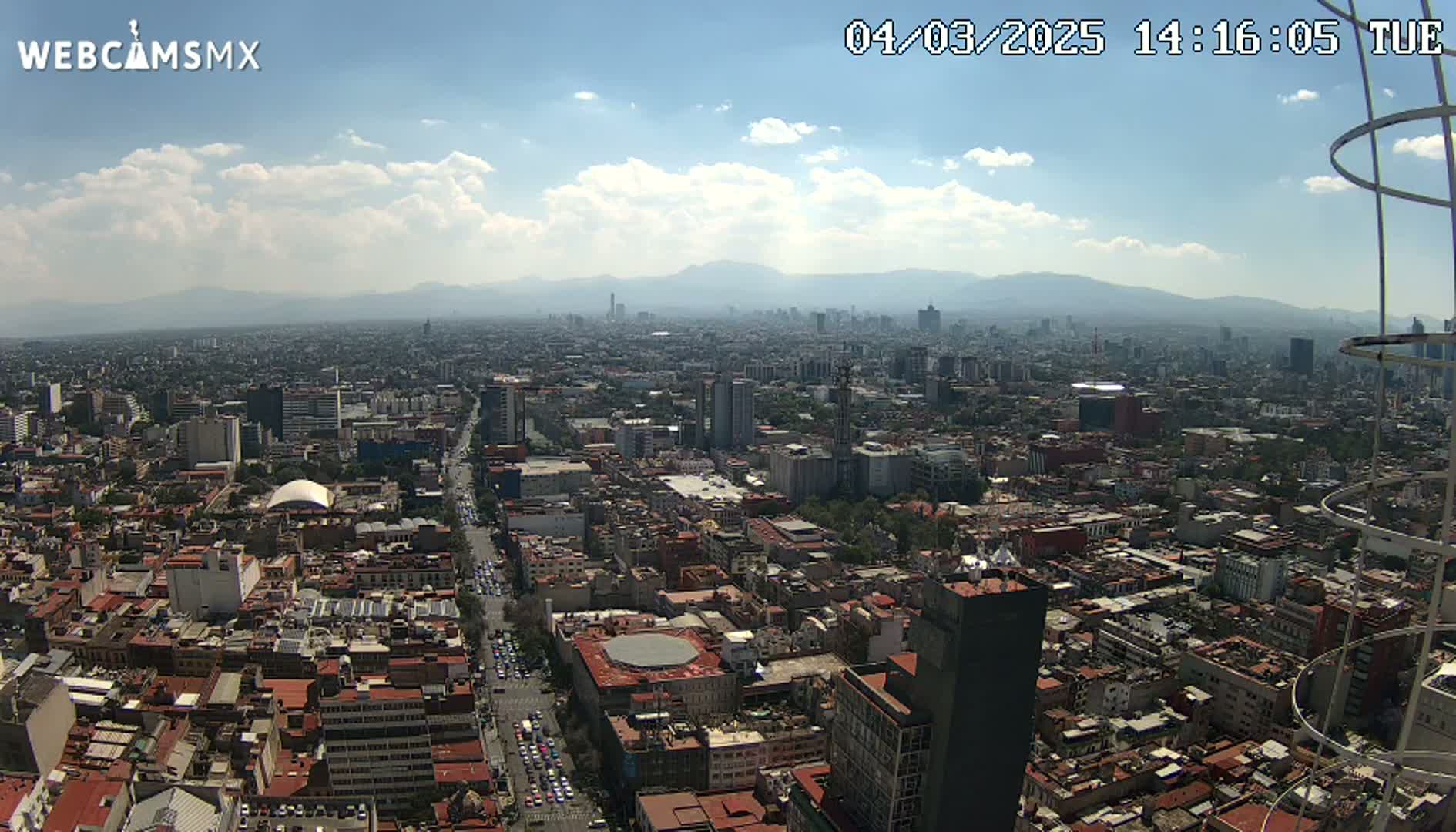 A high-angle, wide shot of a sprawling city under a partly cloudy sky shows a large urban area extending to distant mountains.