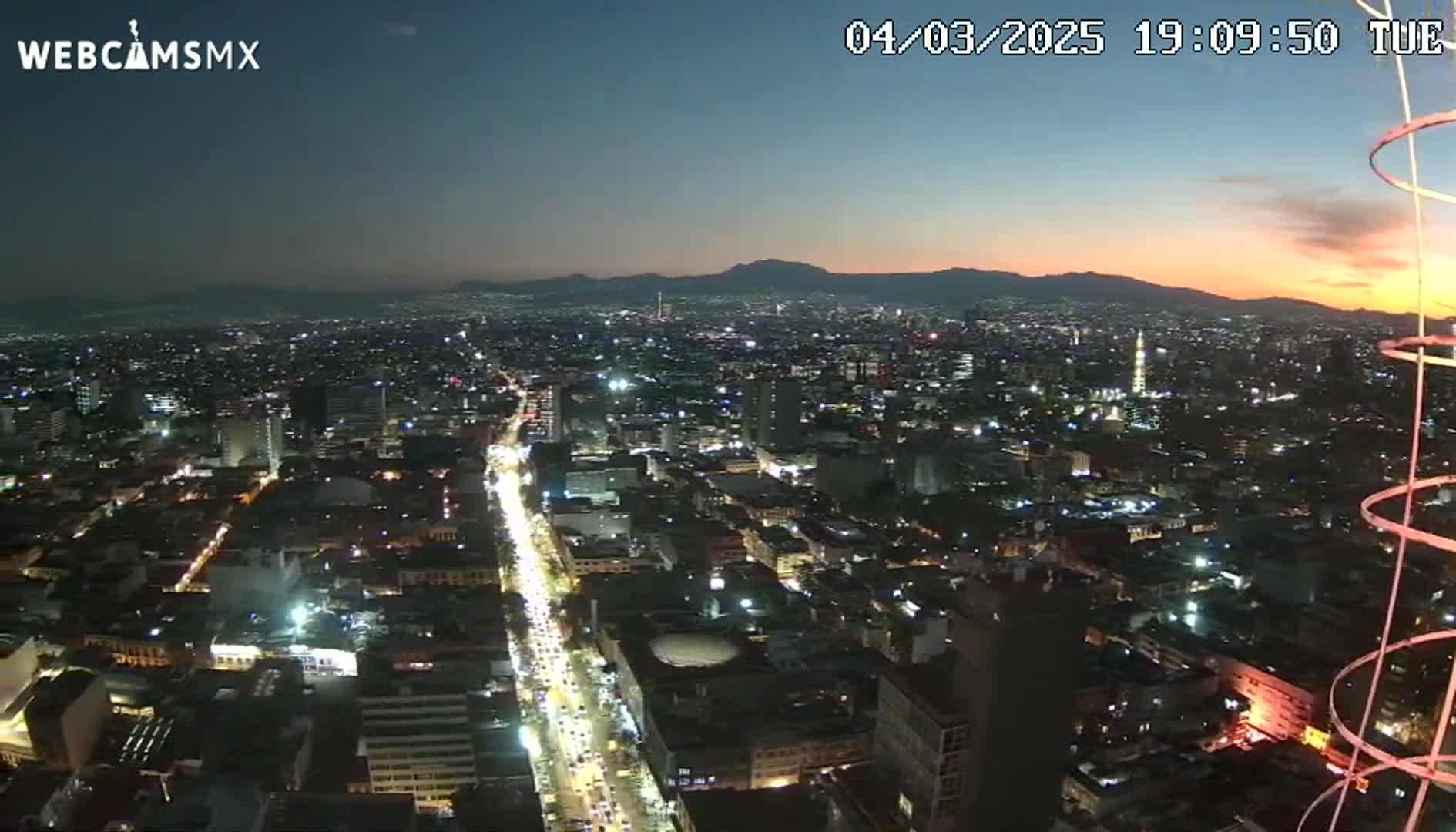 A nighttime, high-angle view of a sprawling city illuminated by streetlights and building lights, with a sunset visible in the distance over a mountain range.