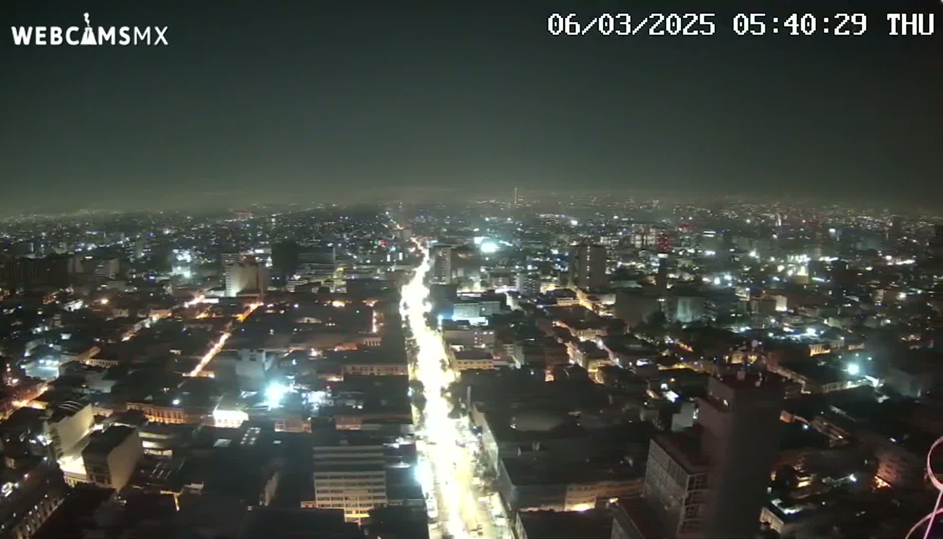 A nighttime aerial view of a city shows a brightly lit main street cutting through a sprawling urban landscape under a hazy sky.