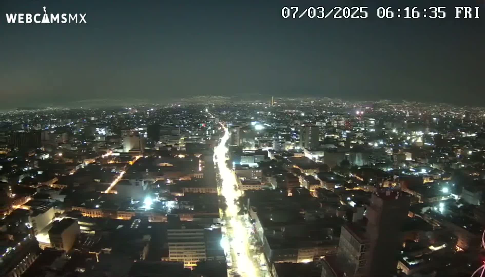 A brightly lit city at night is viewed from above, showing a long, illuminated street stretching through the urban landscape under a clear, dark sky.