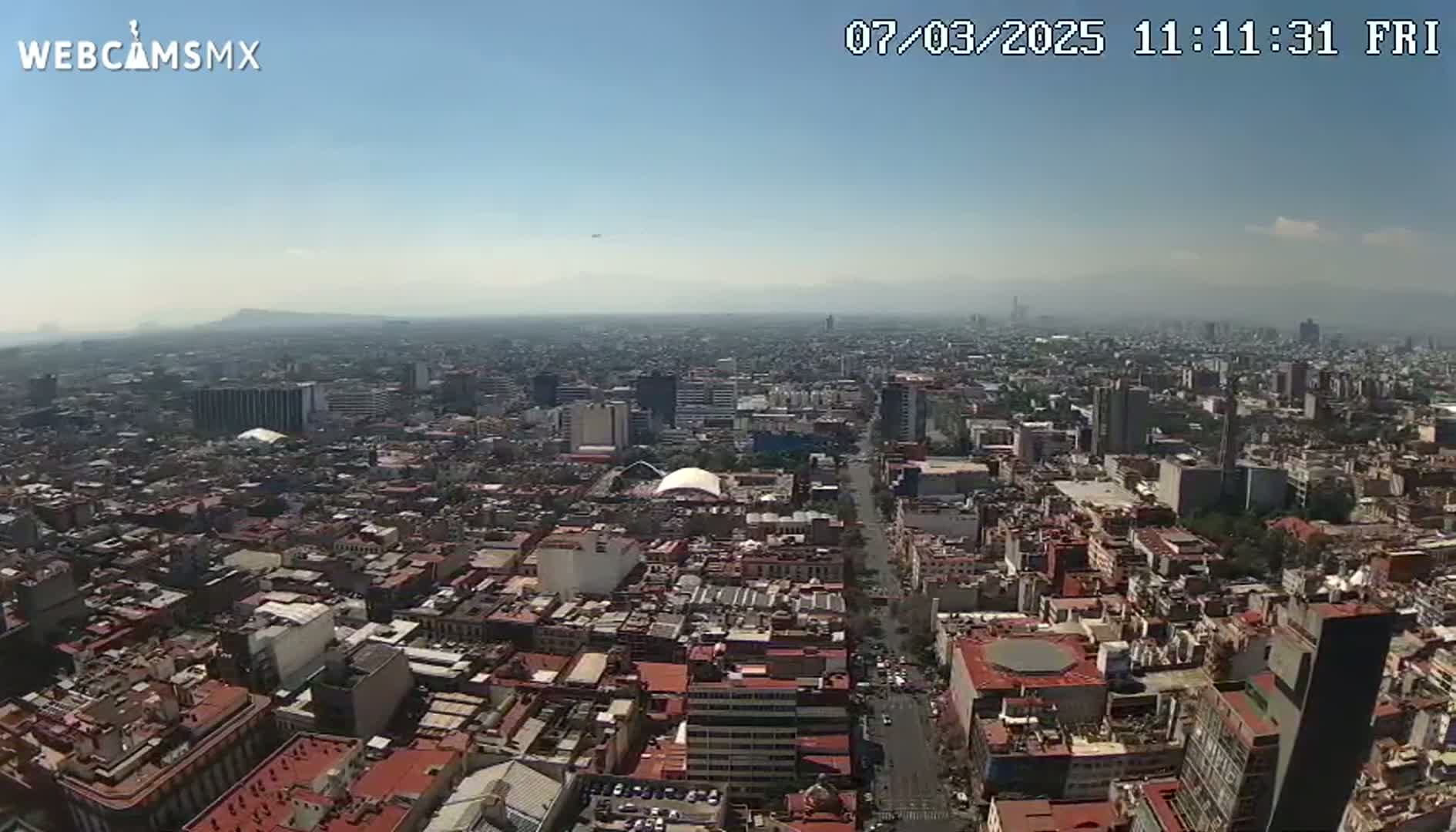 A high-angle, long shot on a clear, hazy day shows a sprawling cityscape with many low-rise buildings, interspersed with taller structures, extending to distant mountains under a pale blue sky.