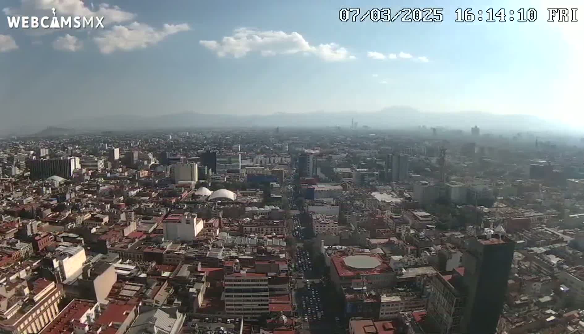 A hazy, sunny day overlooks a sprawling city with numerous buildings, streets, and distant mountains in the background.