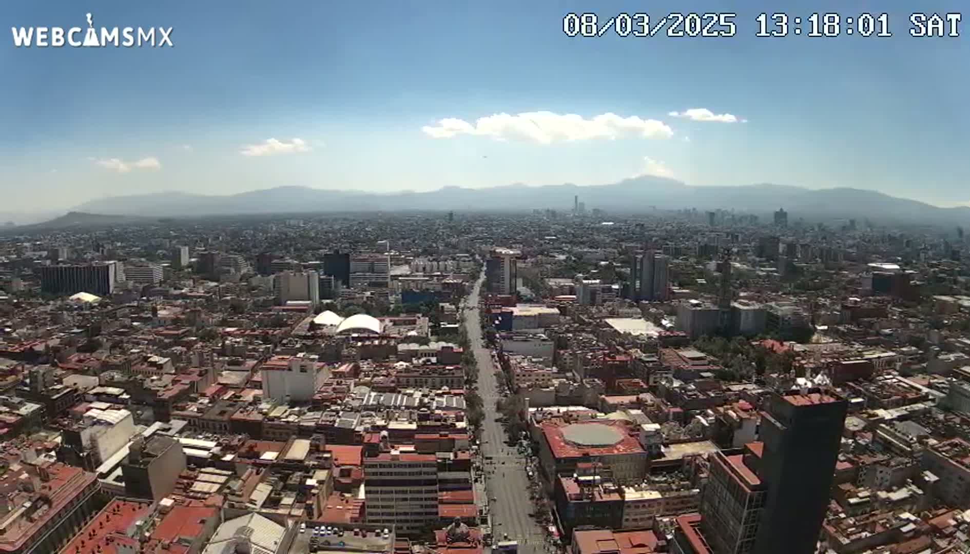 A high-angle view of a sprawling city under a mostly clear sky with a few clouds, mountains visible in the distance.