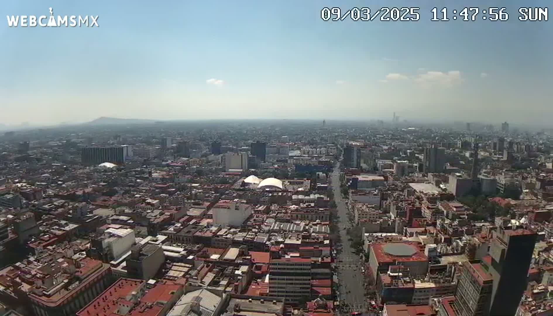 A high-angle, long shot on a sunny day shows a sprawling cityscape extending to hazy mountains in the distance.