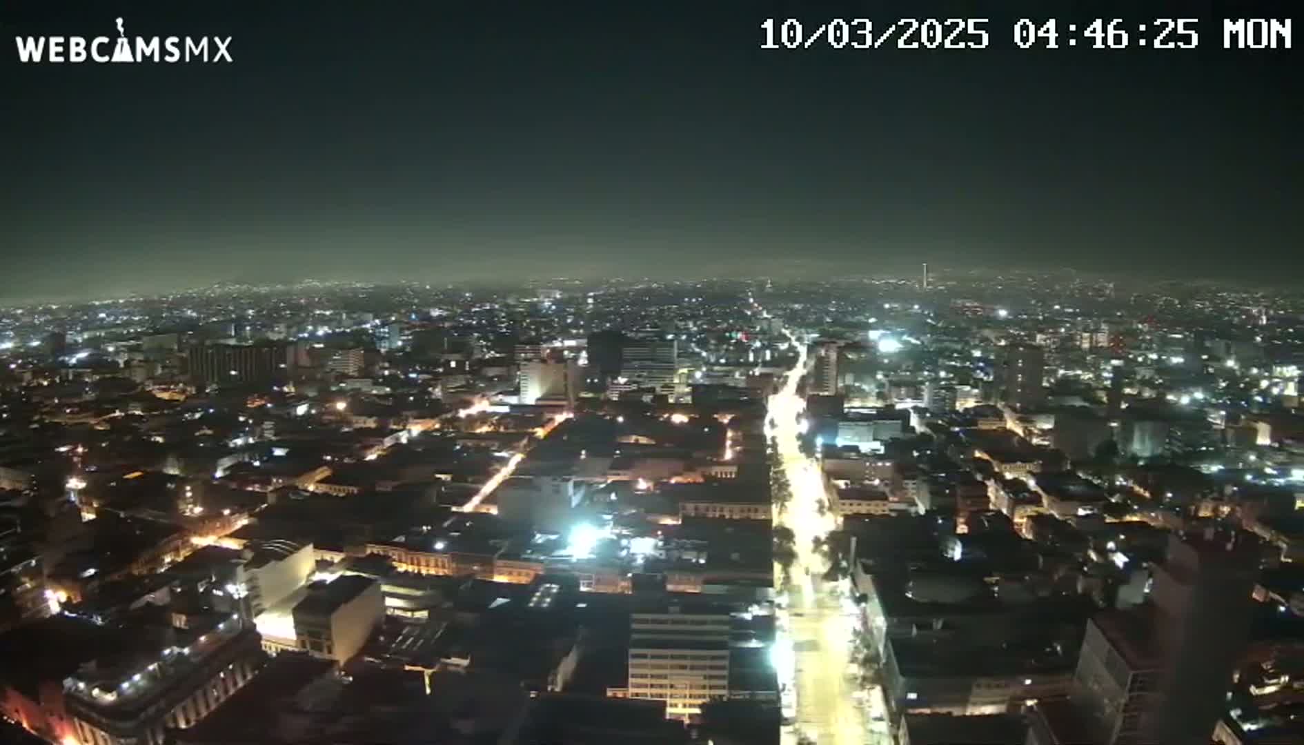 A nighttime aerial view shows a sprawling city with brightly lit streets and buildings under a hazy sky.