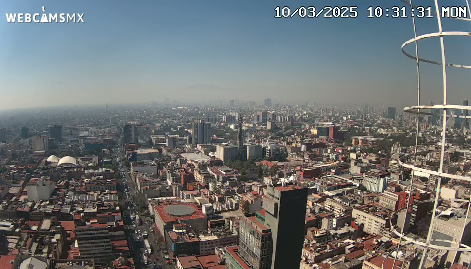 A hazy, sunny day reveals a sprawling cityscape extending to the horizon, viewed from a high vantage point near a metal structure.
