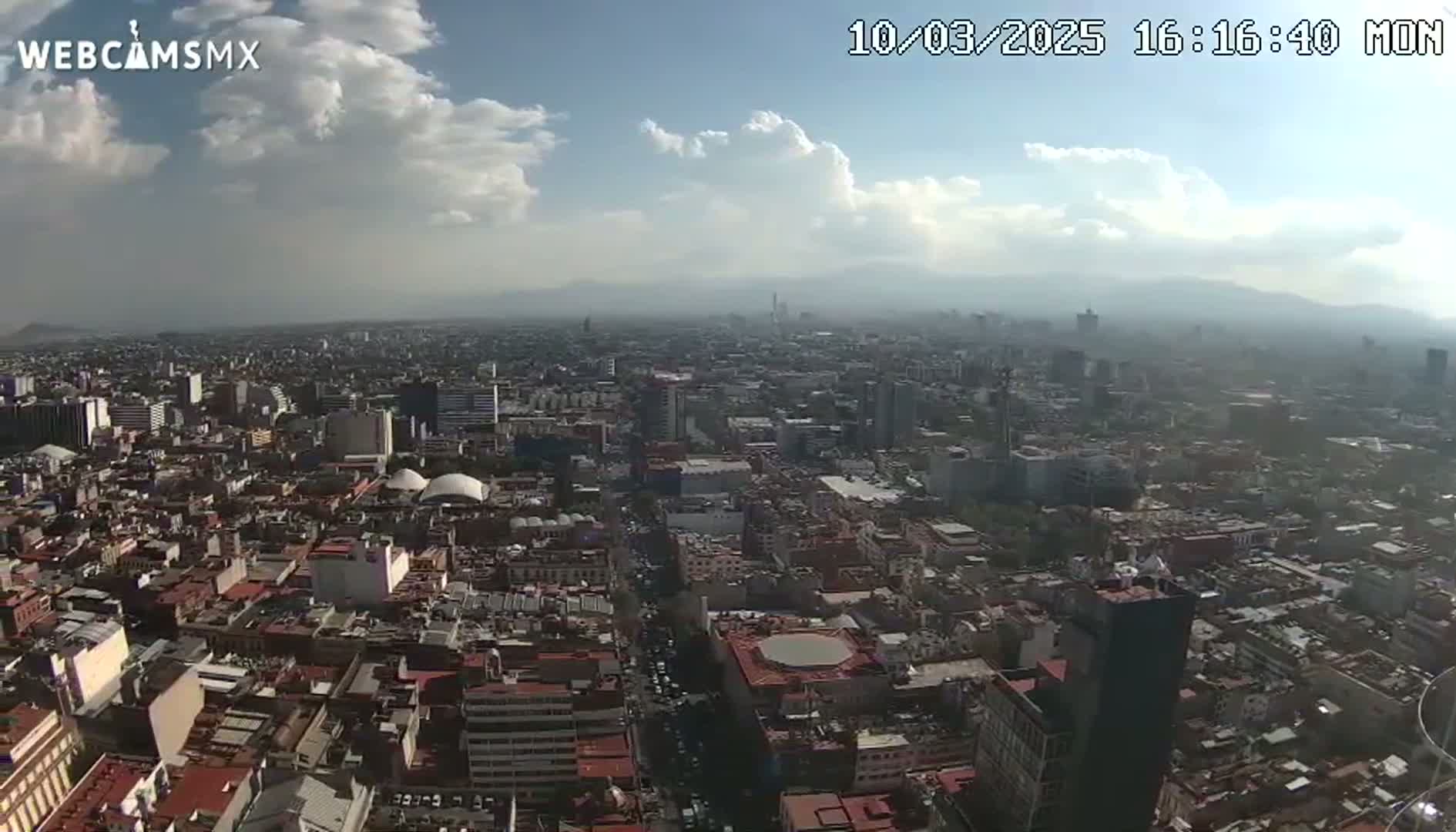A partly cloudy sky overlooks a sprawling city with distant mountains in the background.