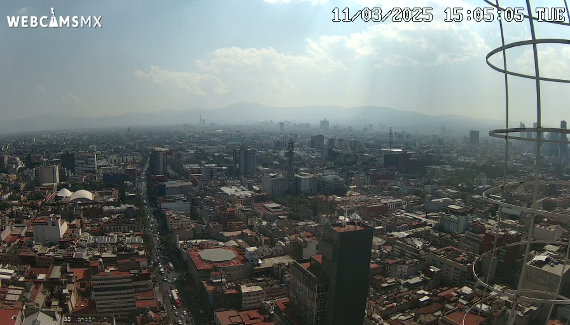 A hazy, partly cloudy day reveals a sprawling cityscape, viewed from a high vantage point, with distant mountains visible on the horizon.