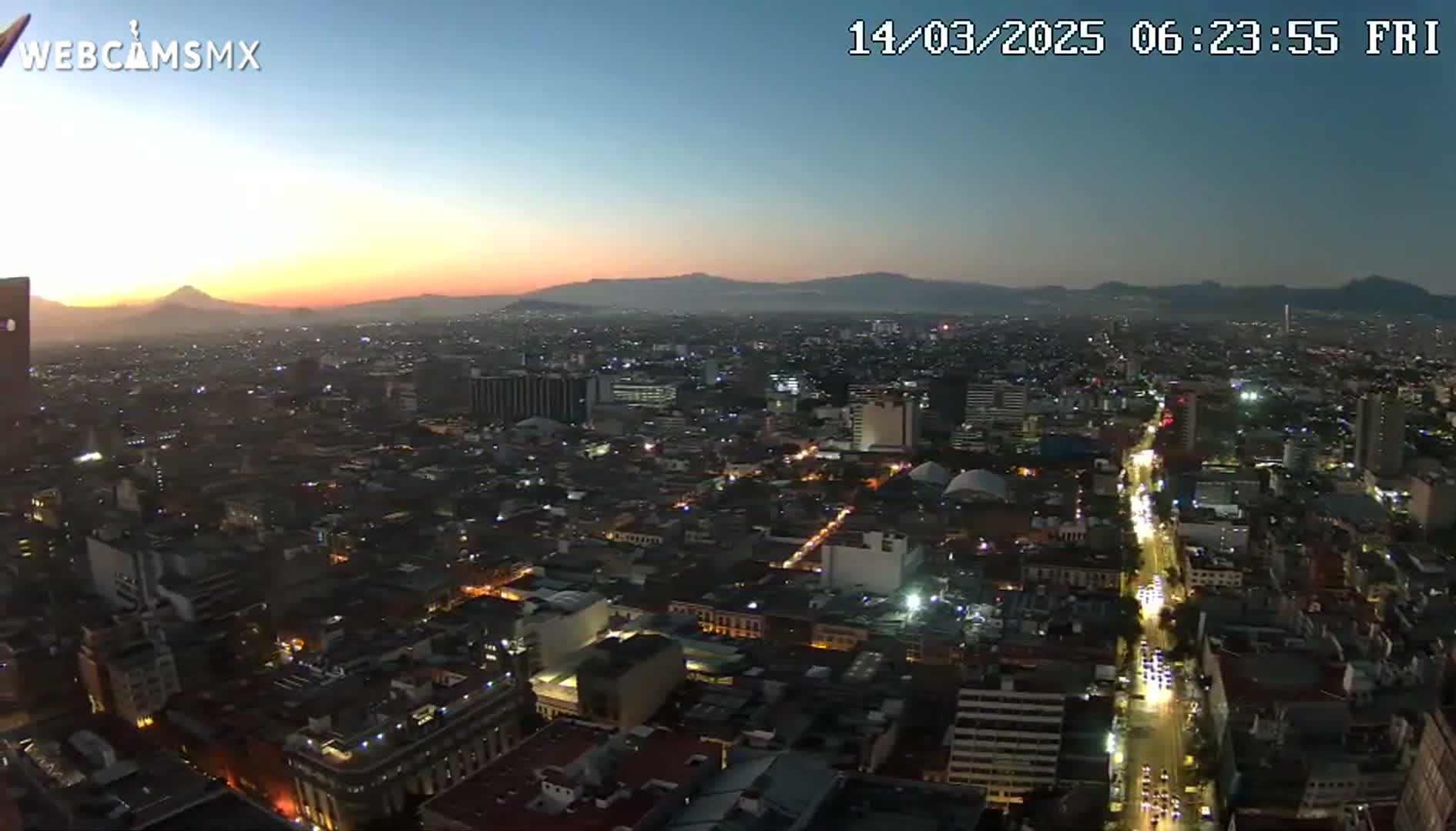 A nighttime, high-angle view of a sprawling city at sunrise, with mountains visible in the distance under a clear sky.