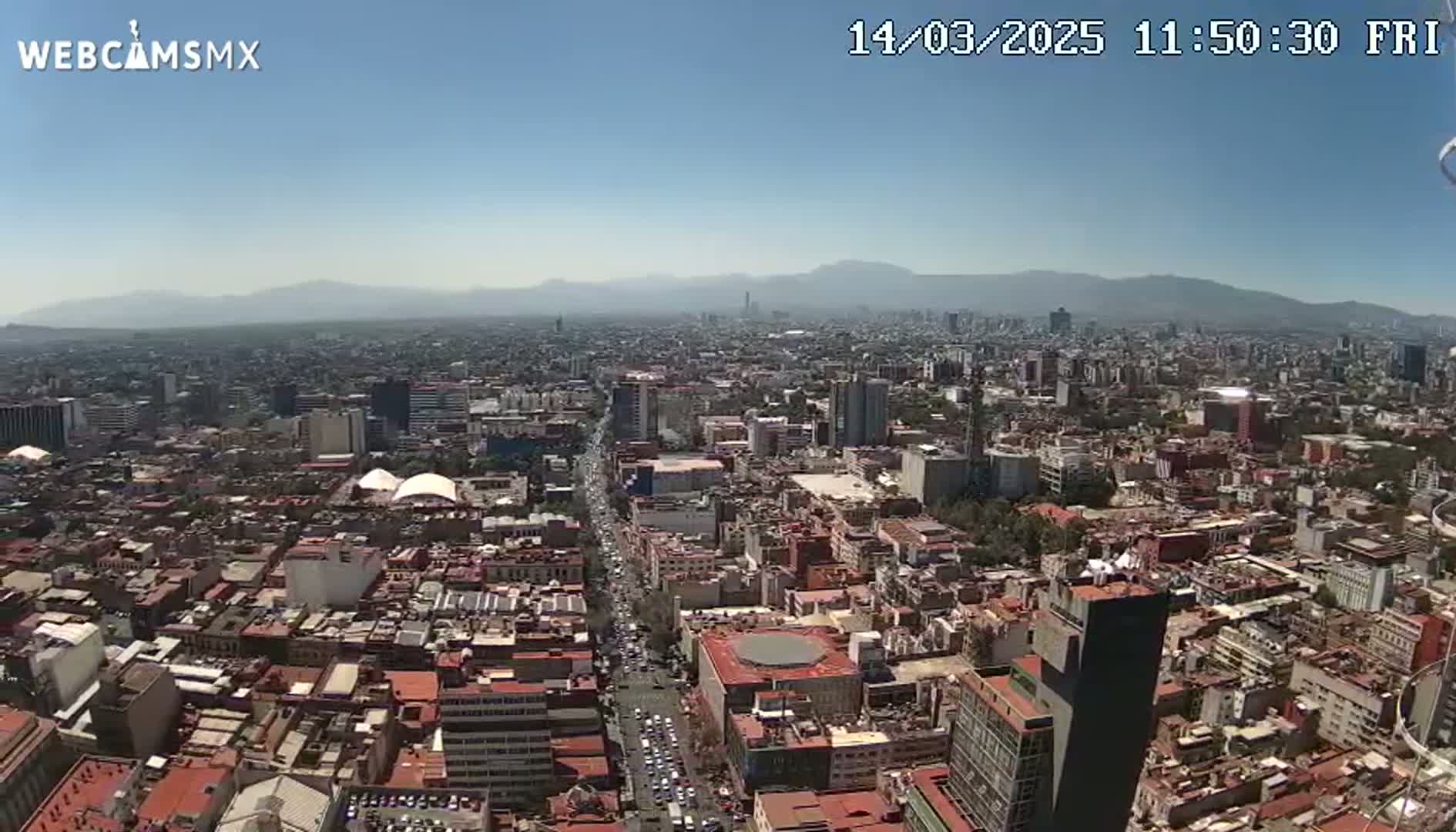 A high-angle, long shot on a clear day shows a sprawling city with many low-rise buildings under a mostly clear sky and distant mountains.