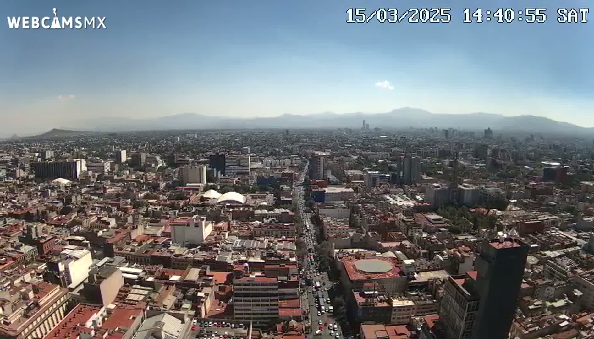 A high-angle, long shot on a clear, sunny day shows a sprawling cityscape extending to distant mountains.