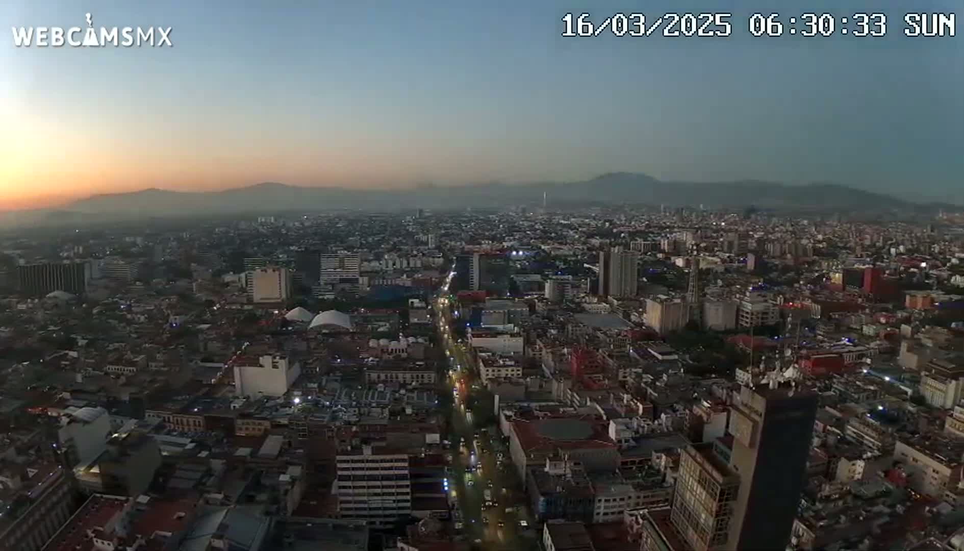 A hazy sunrise illuminates a sprawling city with numerous buildings and streets, mountains visible in the distance.