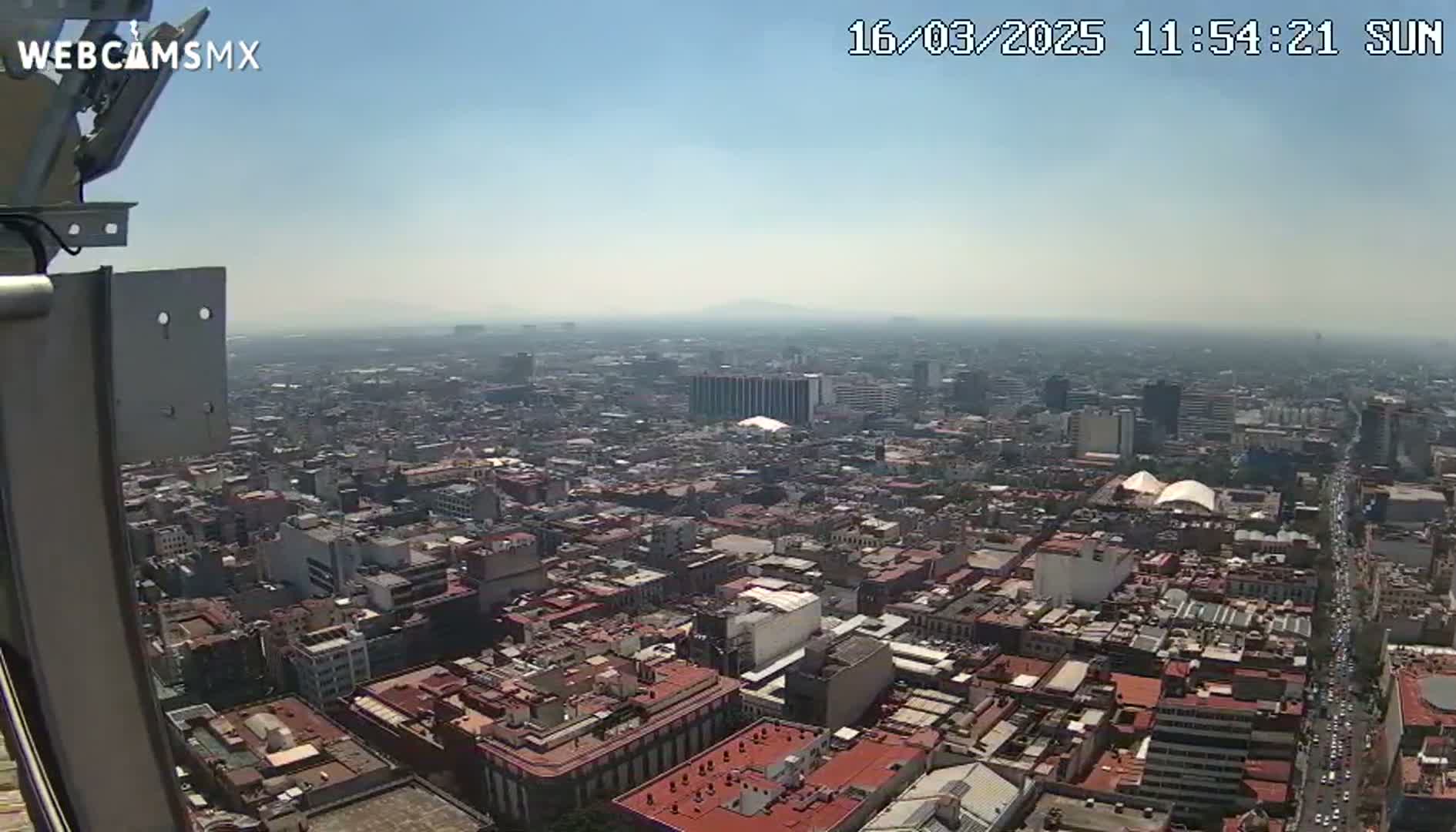 A hazy, sunny day view from a tall building shows a vast expanse of densely packed city buildings extending to the horizon.